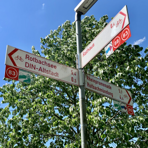 Straßenlaterne vor Baum und blauem Himmel mit drei Wegweisern für Menschen, die zu Fuß oder mit dem Rad unterwegs sind. - Knotenpunktschilder
