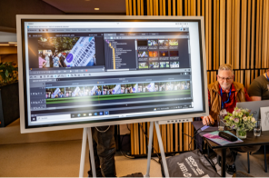 Bildschirm im quer Format, auf dem gezeigt wird, wie ein Film geschnitten wird. Er steht vor einer Wand mit hochkant verlaufenen Brettern im Eingangsfoyer der Kathrin-Türks-Halle Bildschirm im quer Format, auf dem gezeigt wird, wie ein Film geschnitten wird. Er steht vor einer Wand mit hochkant verlaufenen Brettern im Eingangsfoyer der Kathrin-Türks-Halle